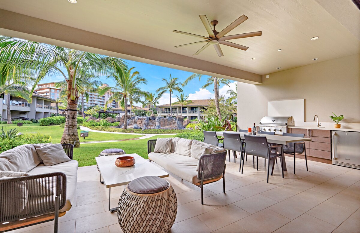 A spacious outdoor lanai offers comfortable seating arranged around a low table, with a built-in BBQ grill and dining area visible. Lush tropical landscaping and palm trees provide a serene backdrop, complemented by a ceiling fan for added comfort.