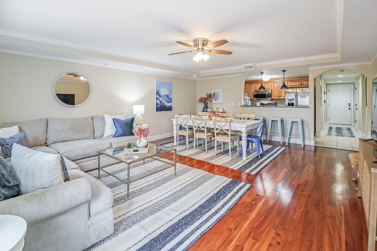 An open-concept living area features a comfortable sectional sofa, complemented by a glass coffee table. A dining table for eight sits nearby, while the kitchen is visible in the background. Warm wood flooring and soft lighting create a welcoming ambiance.