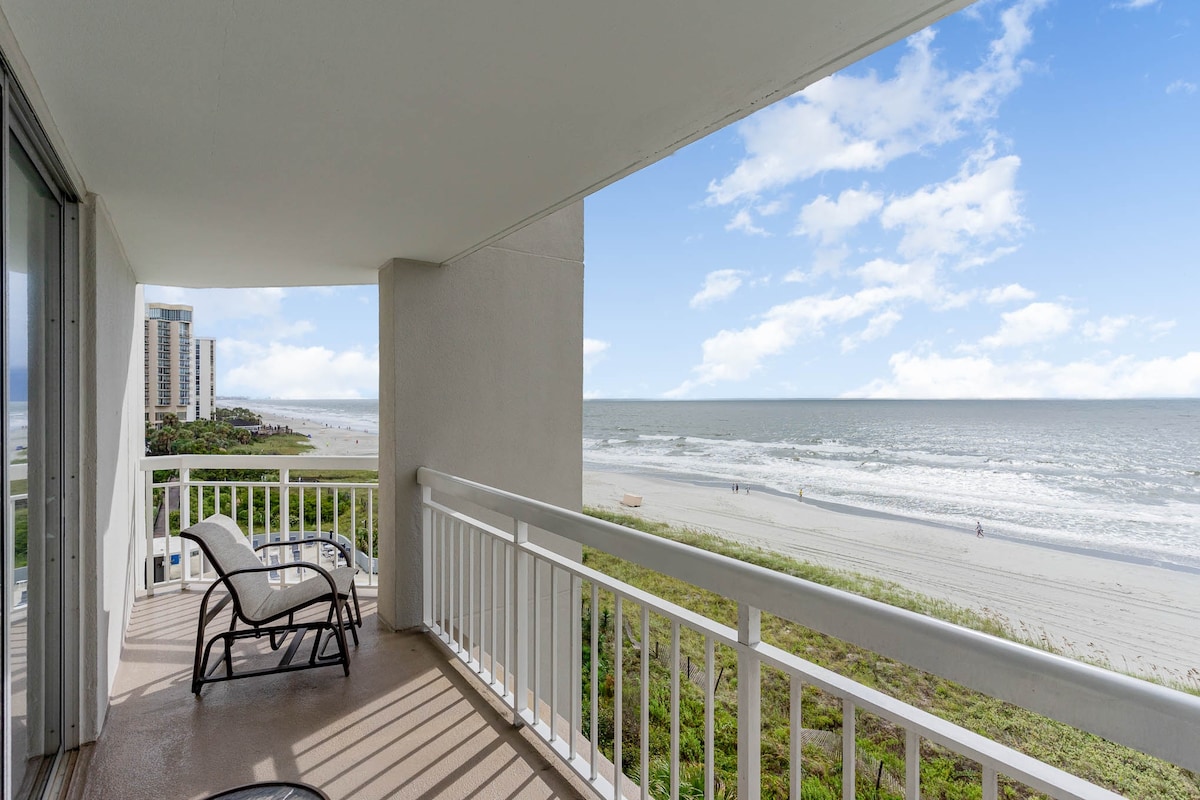 A spacious balcony offers a view of the sandy beach and the Atlantic Ocean. Comfortable seating is positioned to enjoy the refreshing sea breeze. The horizon showcases a blend of blue skies and gentle waves, creating a serene outdoor space for relaxation.
