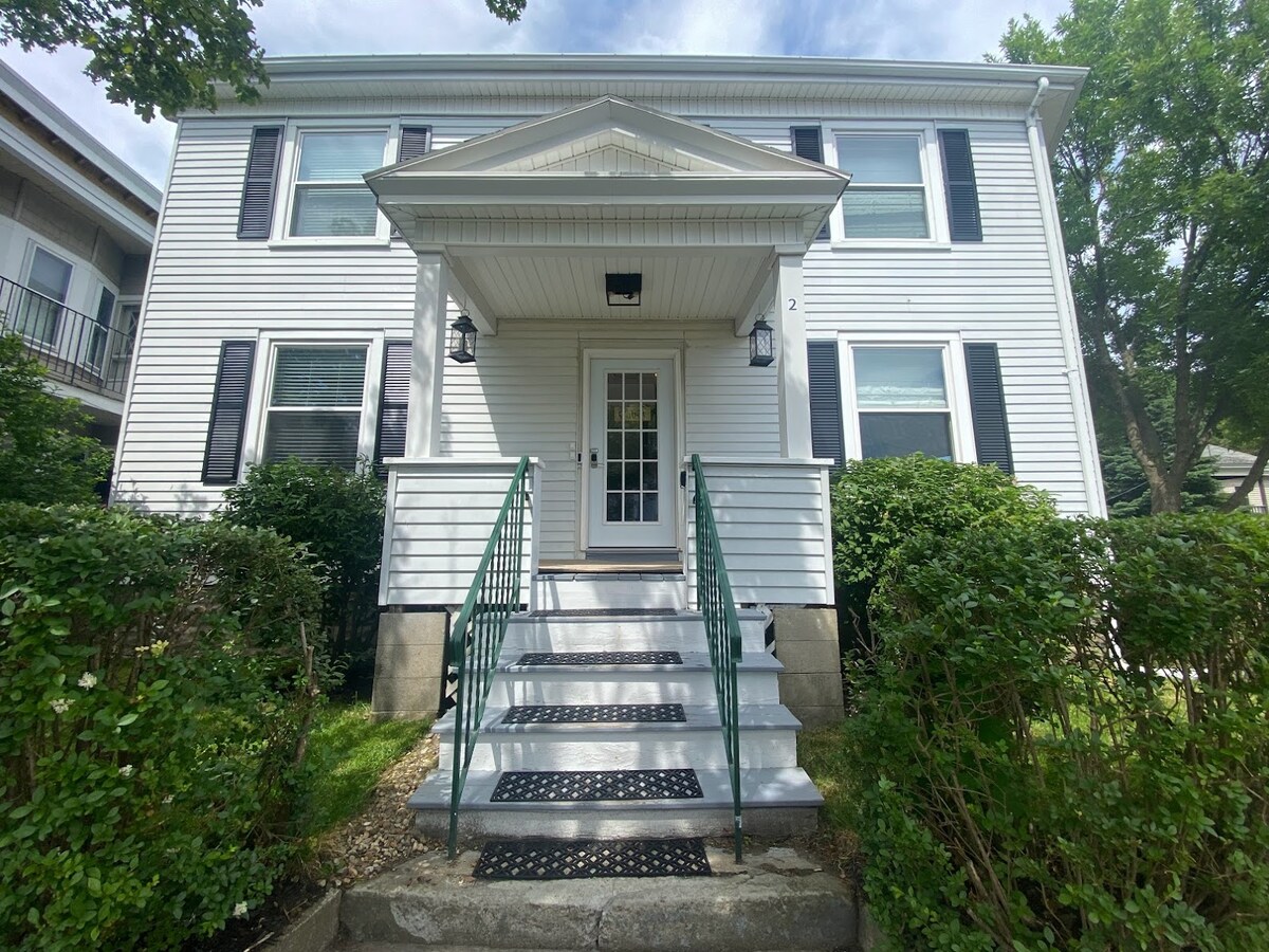 The front exterior of the two-story building is showcased, featuring white siding and black shutters. A set of steps leads to the entry door, flanked by small lanterns. Lush green bushes border the walkway, creating a pleasant foreground.