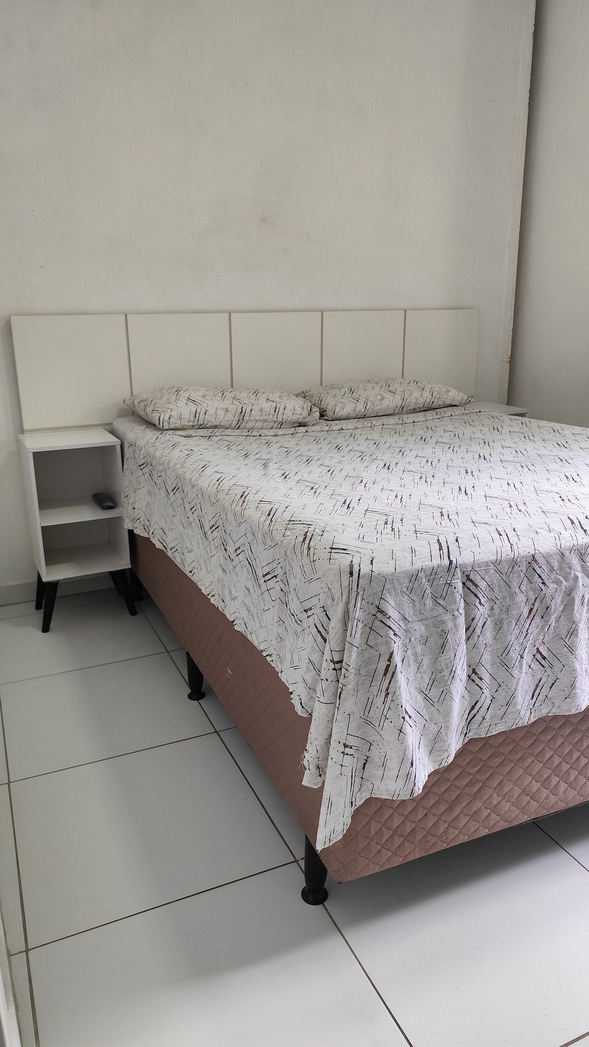 A spacious bed with a patterned white bedspread is positioned against a clean, light-colored wall. Two small pillows rest at the head of the bed. A nightstand with a drawer is placed next to the bed, maintaining a minimalist aesthetic.