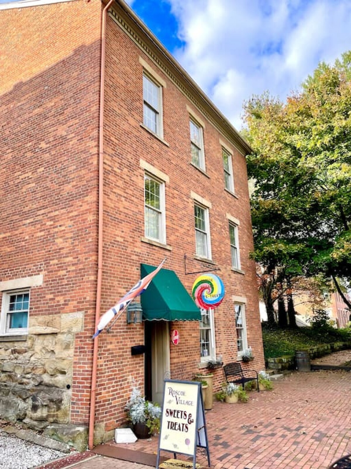 An iconic historic brick building is featured, showcasing large windows that allow natural light to enter. A colorful lollipop sign adorns the entrance, which is framed by a green awning. Outdoor benches are arranged in front, surrounded by landscaped greenery.