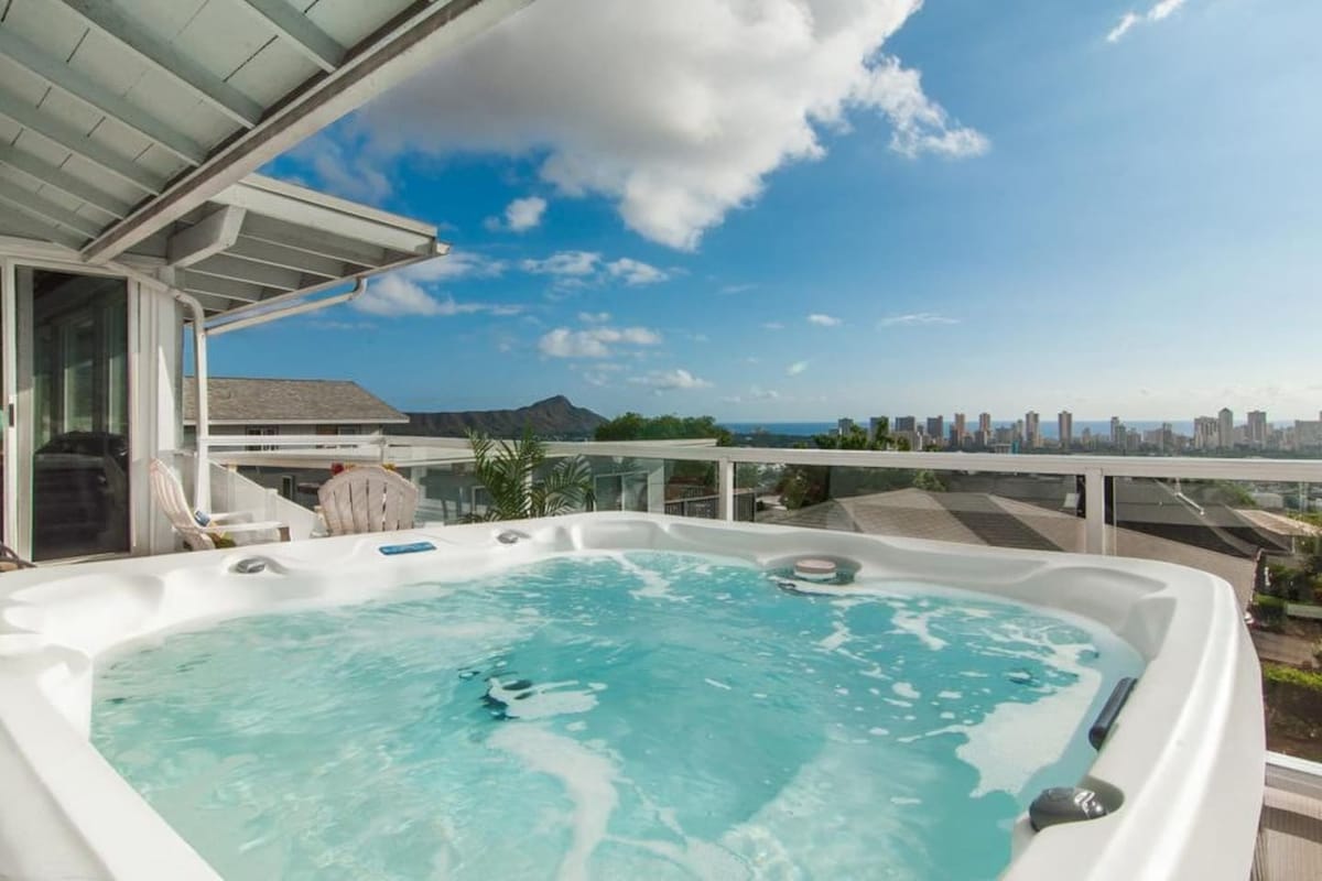 A private hot tub is featured on the balcony, designed for seven people. Clear water glistens under sunlight, with expansive views of the ocean and city skyline visible in the distance. The surrounding area is adorned with tropical foliage, enhancing the serene atmosphere.