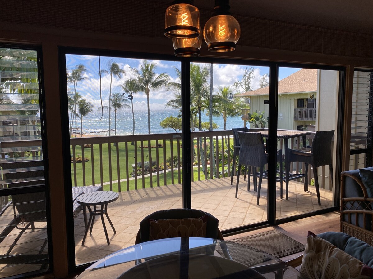 A spacious lanai is visible through expansive glass doors, offering a view of the ocean and palm trees swaying in the breeze. Outdoor seating is arranged at a high-top table, complementing the inviting interior ambiance of the condo.