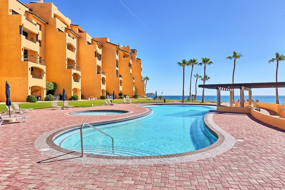 A sparkling pool is framed by a landscaped area with palm trees and vibrant orange buildings. The pool boasts a curvy shape and steps leading into the water. Lounge chairs are arranged around the poolside, with a shaded seating area visible in the background.