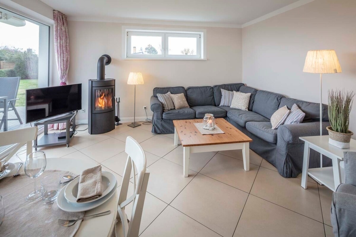 A comfortable living area features a spacious sectional sofa with decorative cushions, a wooden coffee table, and a flat-screen television. Natural light enters through large windows, while a wood-burning stove provides warmth. A dining table is set with cutlery and plates, inviting a shared meal.