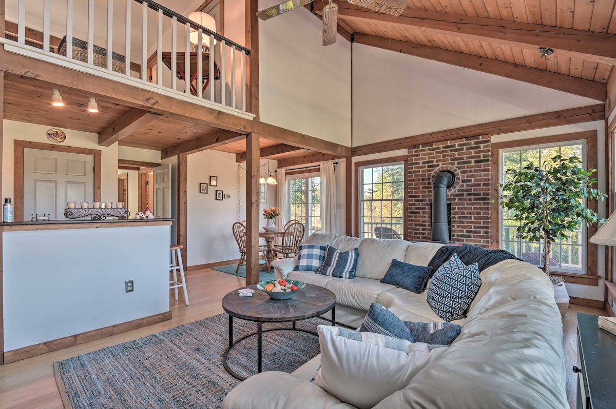 An open living area features a large sectional sofa, a round coffee table, and a rustic wood-burning stove. Natural light fills the space through multiple windows, and a wood-paneled ceiling enhances the cozy ambiance. A dining area and kitchen bar are visible in the background.