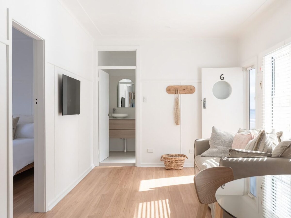 A bright and airy living space features light wooden flooring and large windows allowing natural light to fill the room. A comfortable sofa and a dining table are visible, with a bathroom area seen through an open doorway. Soft, neutral decor accents create a serene environment.