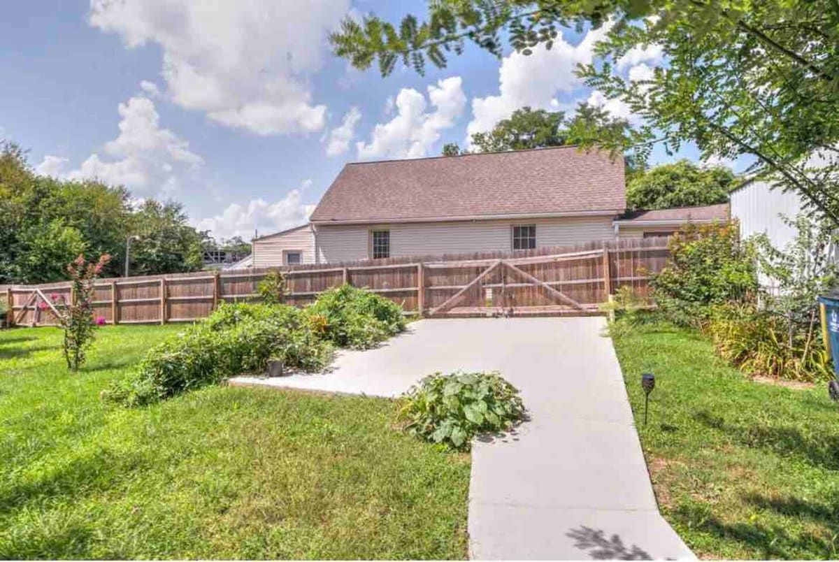 The spacious backyard is surrounded by a tall wooden fence, enhancing privacy. A paved pathway leads to a gate, bordered by vibrant greenery and plants. Bright blue skies and fluffy clouds create a soft backdrop over the serene outdoor space.