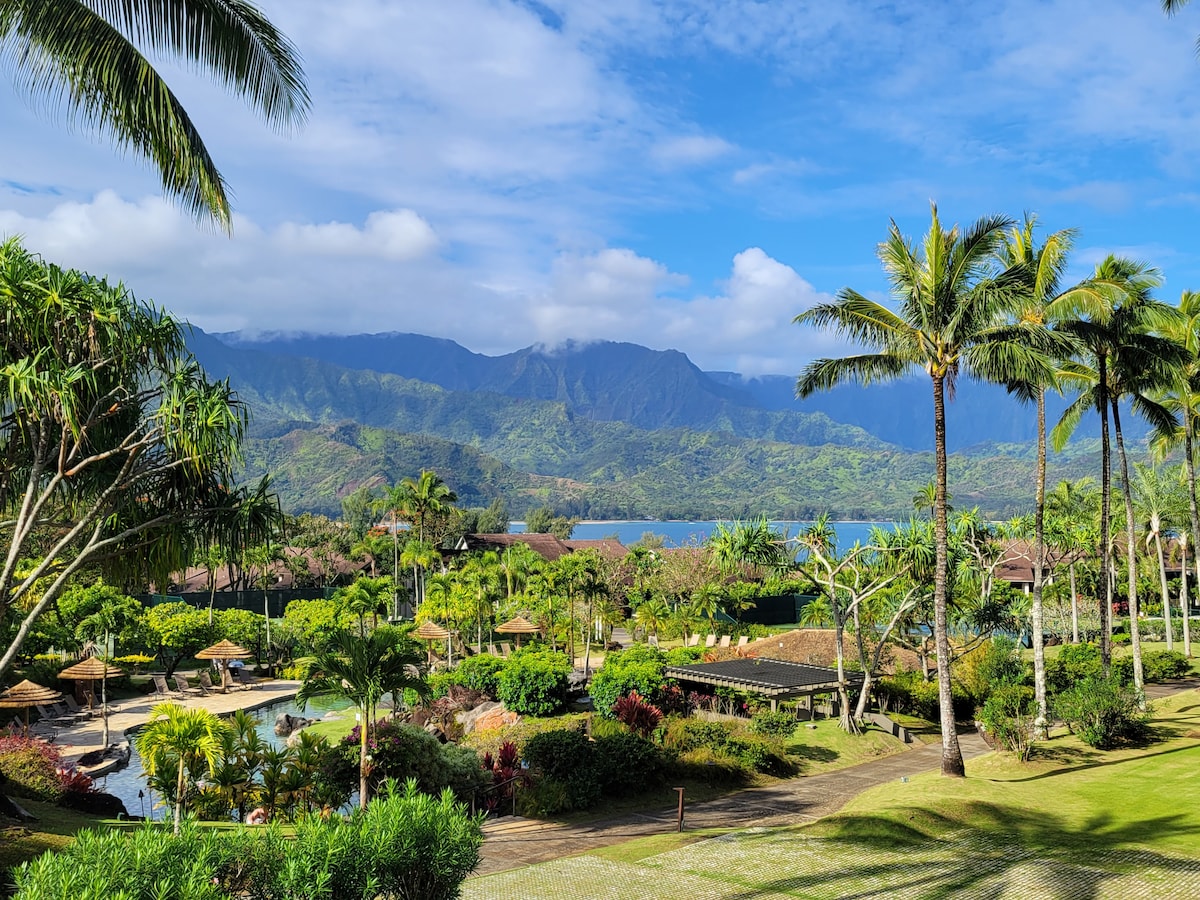 The image showcases a lush tropical landscape with vibrant palm trees and manicured gardens. Majestic mountains rise in the background, overlooking Hanalei Bay. The scene includes tranquil water features and distant views of the ocean beneath a bright blue sky, creating a serene atmosphere.