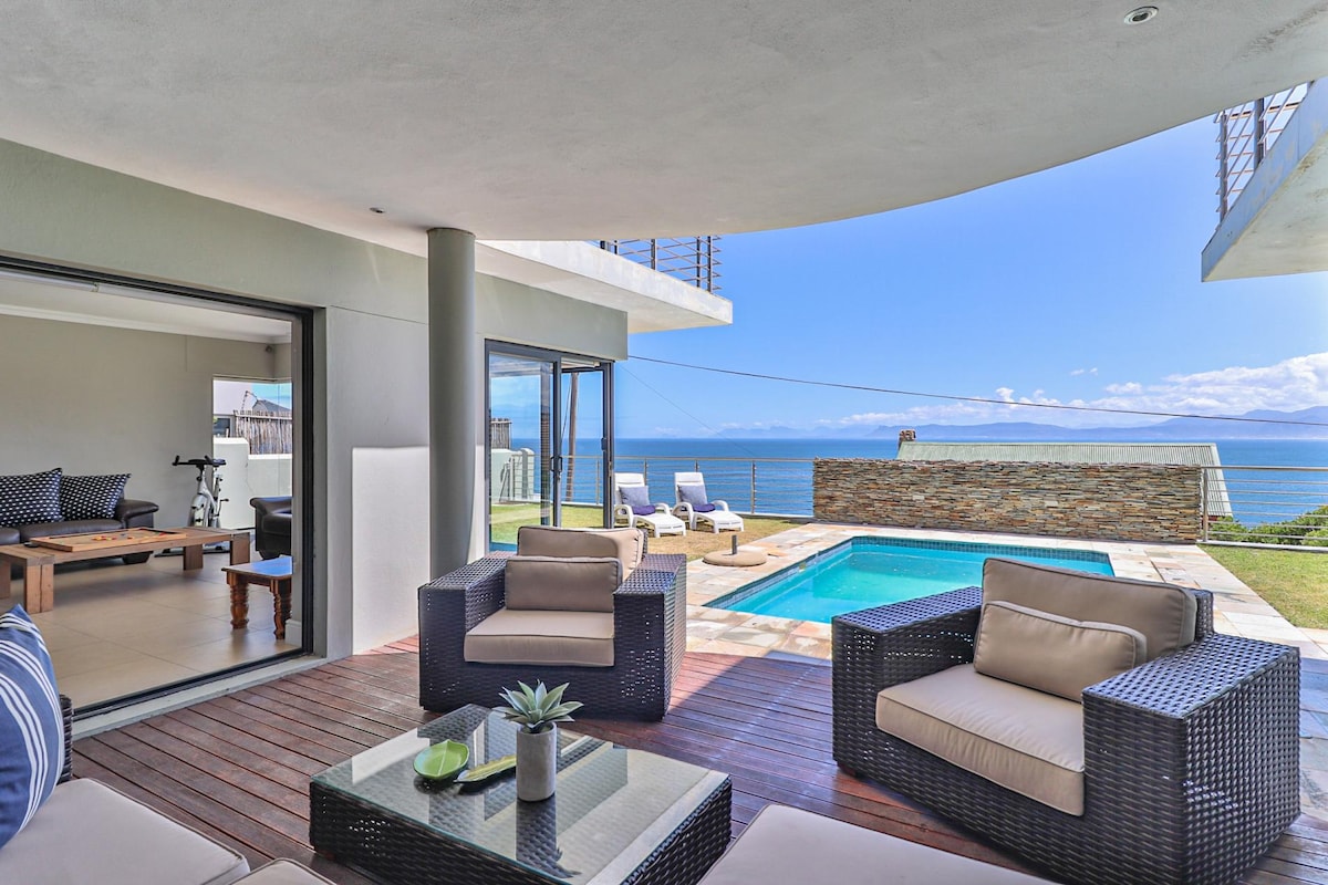 An outdoor lounge area is showcased, featuring comfortable seating and a glass-topped table. The space opens to a swimming pool, with sun loungers positioned nearby, all overlooking the expansive ocean view and clear blue sky.