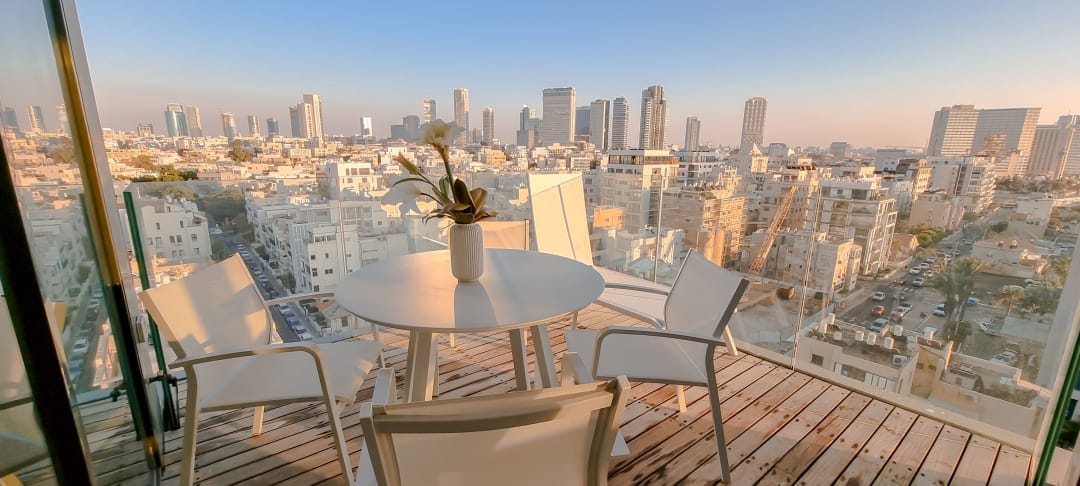 A spacious balcony is shown with modern white furniture, including a circular dining table and four chairs. A potted orchid centerpiece sits on the table, with a panoramic view of the city skyline in the background, showcasing a blend of architectural styles.