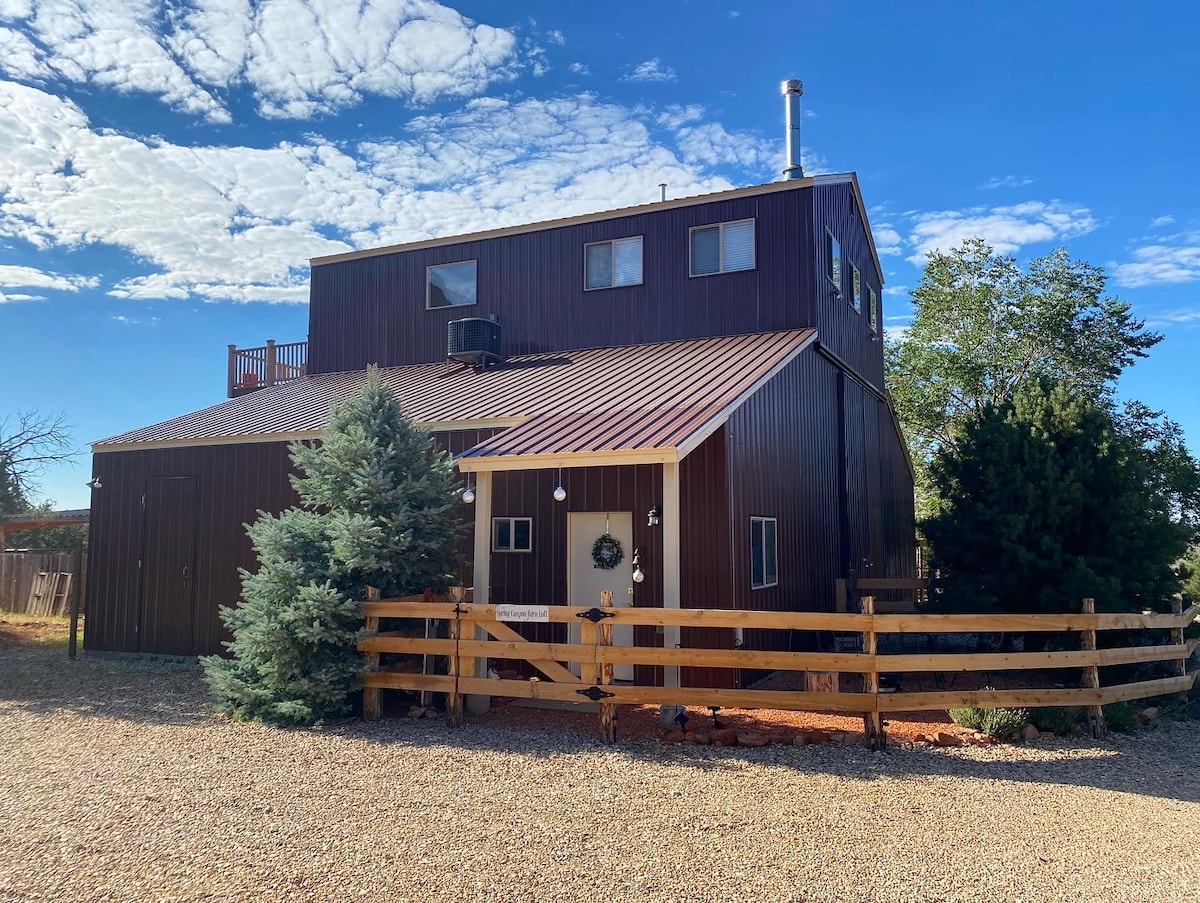 The exterior of the Barn Loft is shown, featuring a two-story brown structure with a metal roof. A wooden fence encircles the property, complemented by green shrubs and trees. The sky is clear, with scattered clouds overhead, creating a peaceful atmosphere.
