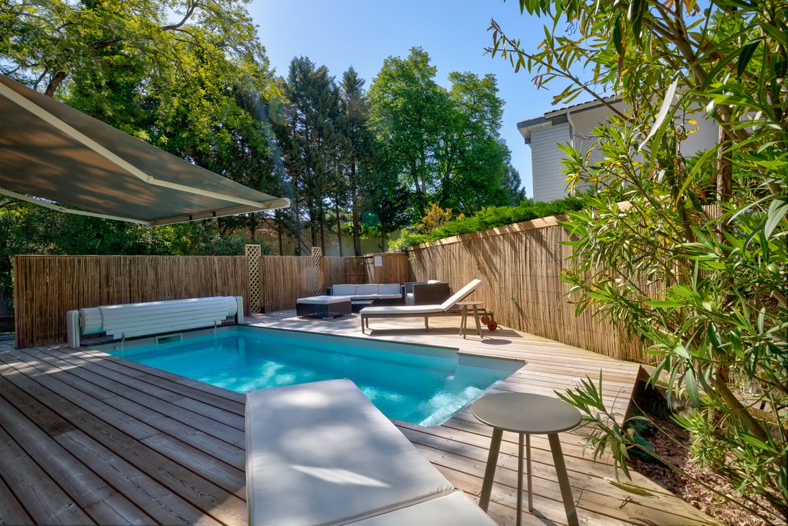 A private pool area is presented featuring a rectangular pool surrounded by wooden decking. Lounge chairs are positioned near the water, with a shaded seating area providing a space for relaxation amid lush greenery and bamboo fencing, ensuring a tranquil outdoor environment.