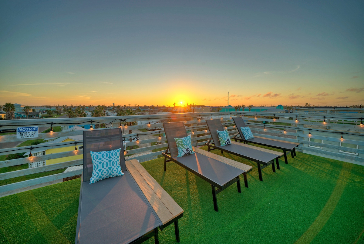 The rooftop deck features modern loungers arranged for relaxation, with vibrant pillows enhancing comfort. A captivating sunrise casts a warm glow over the surroundings, creating an inviting atmosphere. Twinkling lights are visible in the background, adding to the tranquil coastal vibe.