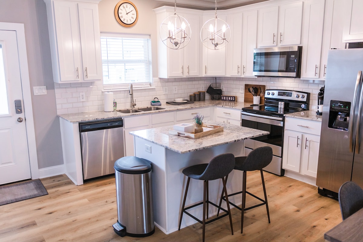 A modern kitchen features white cabinetry and a marble countertop island with two bar stools. Stainless steel appliances include a refrigerator, dishwasher, and oven. Ambient lighting is provided by decorative pendant fixtures, and a clock is mounted on the wall above the sink area.