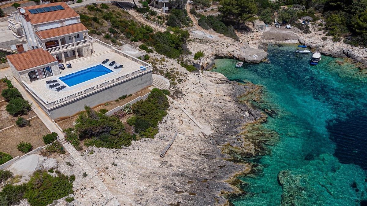 An aerial view captures a spacious holiday home with a large pool surrounded by sun loungers. The property is set against a backdrop of clear turquoise waters and rocky coastline, highlighting its seafront location near the beach.