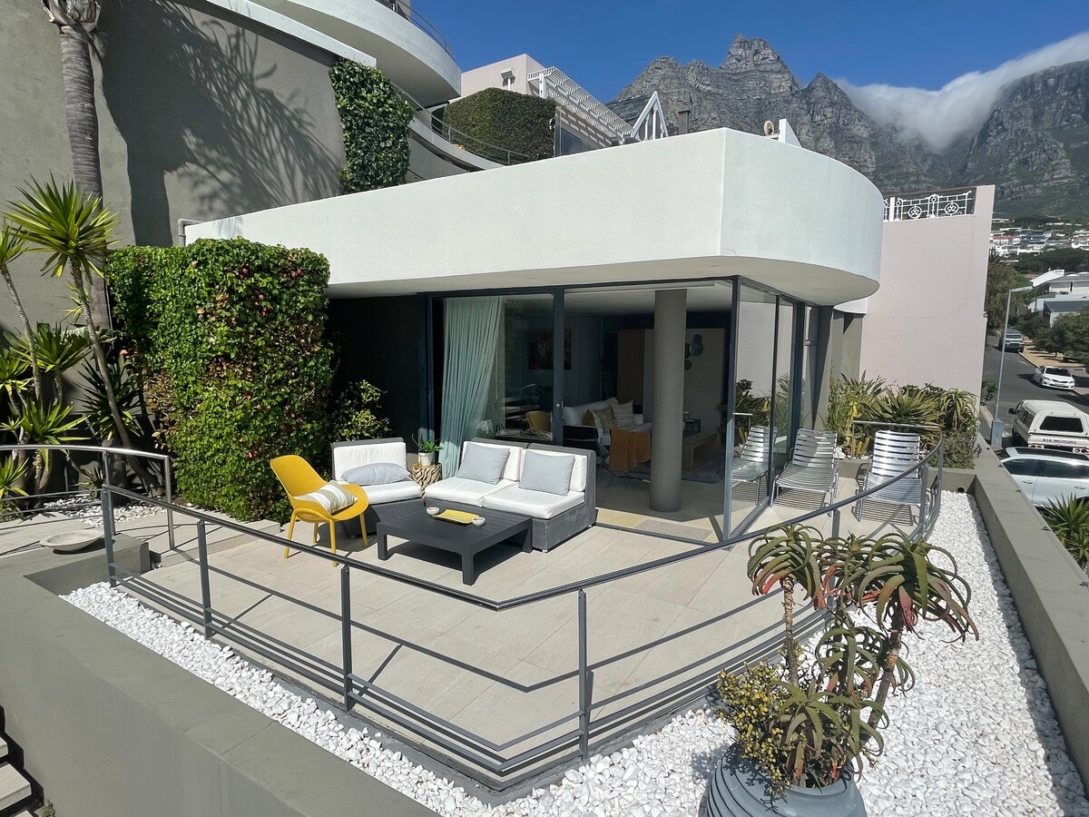 A modern wrap-around patio is surrounded by lush greenery, showcasing comfortable outdoor seating arrangements. Glass sliding doors allow for easy access to the interior space, where contemporary furnishings are visible. The backdrop features the scenic Table Mountain, creating a serene setting.
