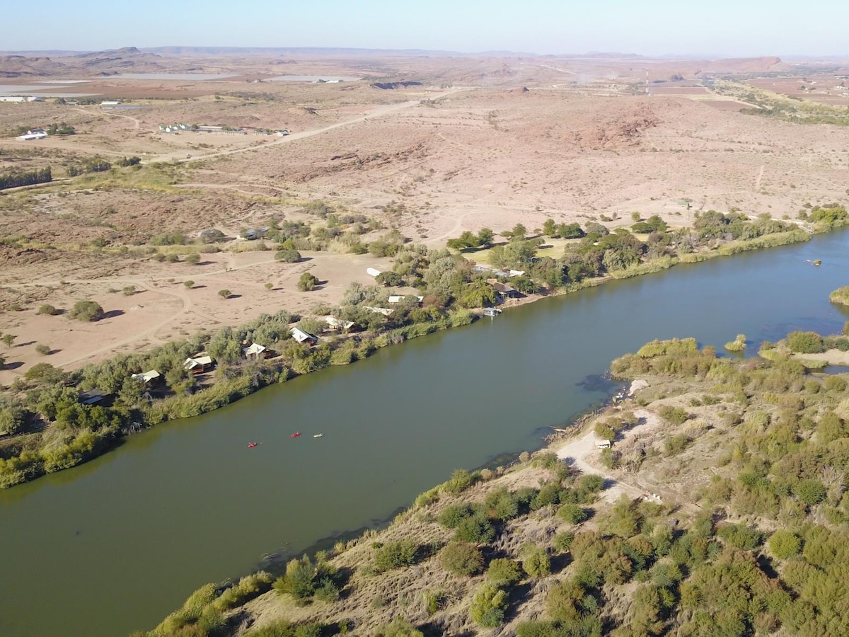 A serene river meanders alongside a landscape characterized by dense greenery and arid terrain. The image captures several cabins nestled by the water's edge, with a clear view of the surrounding hills and a few kayaks on the river, inviting outdoor exploration.
