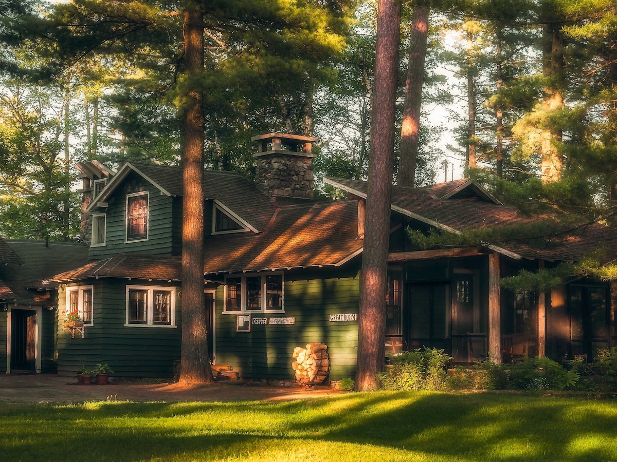 The exterior of a rustic cabin is framed by tall pine trees. Sunlight filters through the branches, creating warm shadows on the green wooden facade. A stone chimney is visible, and a stack of firewood is neatly arranged beside the entrance.