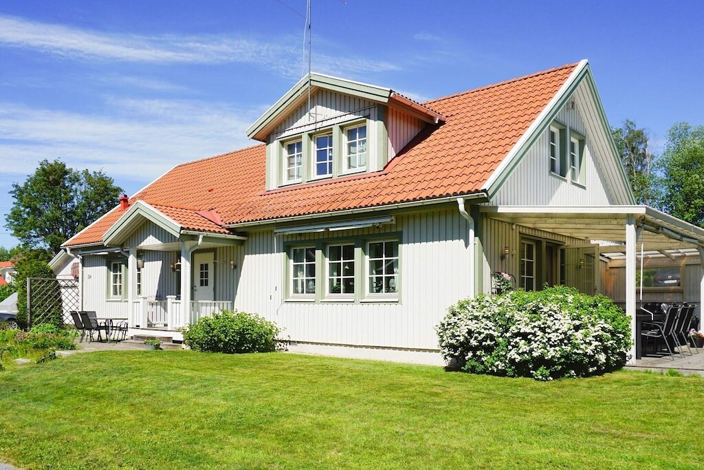 A two-story villa features a vibrant red-roofed exterior with large windows, surrounded by a well-maintained garden. A covered terrace extends from the house, offering outdoor seating options. The lawn is expansive, providing a spacious area for relaxation and enjoyment.