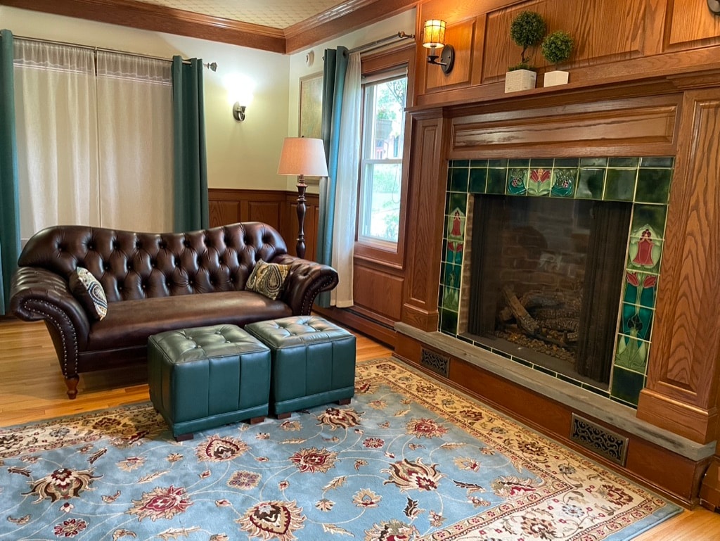 A cozy living area features a brown leather chesterfield sofa, complemented by teal upholstered ottomans. A decorative fireplace, adorned with colorful tiles, adds character to the space, while large windows draped with beige curtains invite natural light.