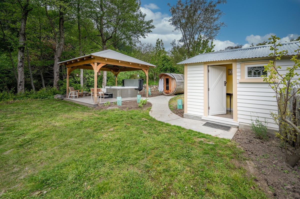 An outdoor area is presented, featuring a covered structure with seating, a sheltered spa pool, and a sauna. A concrete path leads to two individual buildings, surrounded by green lawn and trees, creating an inviting and private space for relaxation.