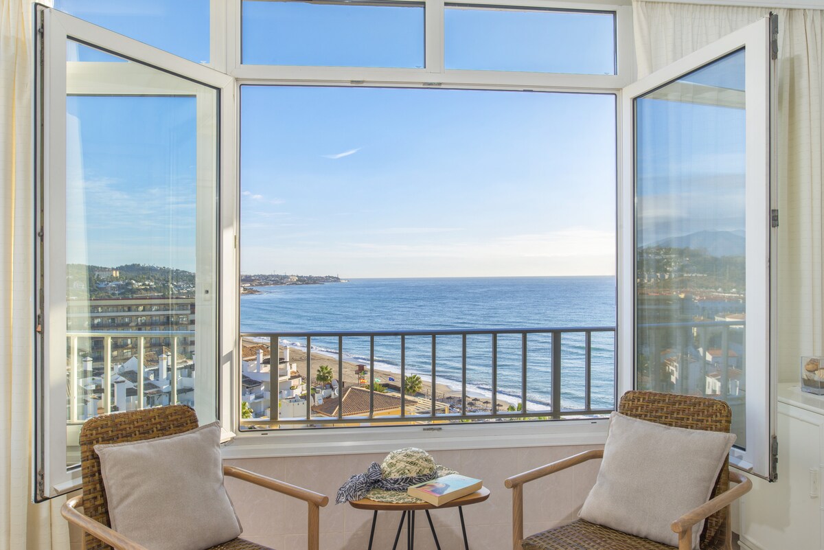 Large windows frame a scenic view of the sea and coastline from a cozy nook. Two armchairs with soft cushions are placed beside a small table, inviting relaxation while enjoying the natural light and expansive ocean vista.