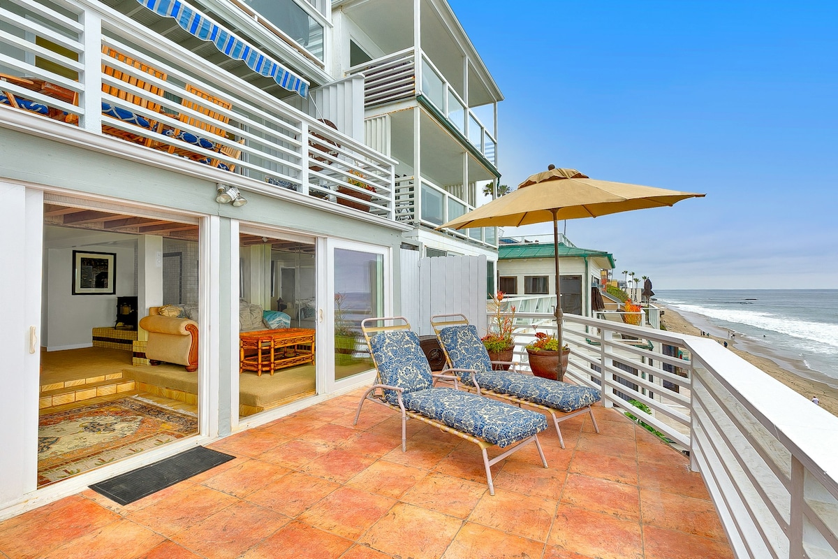 A private outdoor deck is shown with two blue-patterned lounge chairs positioned under a large umbrella. The view extends toward the coastline, with waves gently lapping at the sandy beach. Glass doors connect the outdoor space to the interior living area.