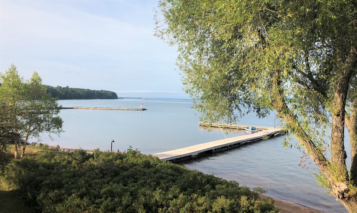 A tranquil view of Lake Superior showcases a shared dock extending into the calm waters. Lush greenery lines the shore, while a distant pier is visible on the horizon. The scene offers a serene atmosphere with soft lighting reflecting off the water.