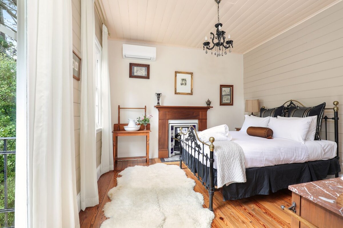 The bedroom features a spacious bed with white linens and a decorative black iron bed frame. A wooden bedside table holds a decorative bowl, while an elegant chandelier hangs above. Natural light pours in through the windows, highlighting the warm wooden flooring and soft area rug.