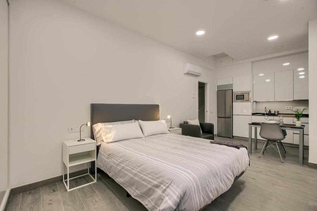 A well-appointed studio apartment features a spacious bed with a striped blanket and a bedside table with a lamp. A kitchenette with modern appliances is visible in the background, along with a dining area that includes a table and chairs.