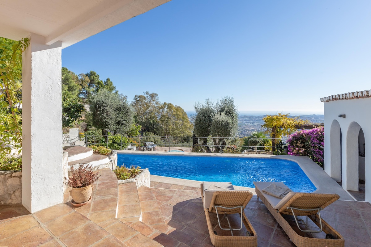 An inviting outdoor space features a shimmering pool surrounded by terracotta tiles. Two lounge chairs are positioned near the water, offering a relaxing view of the lush landscape and distant sea. Vibrant plants and trees frame the scene, enhancing the serene environment.