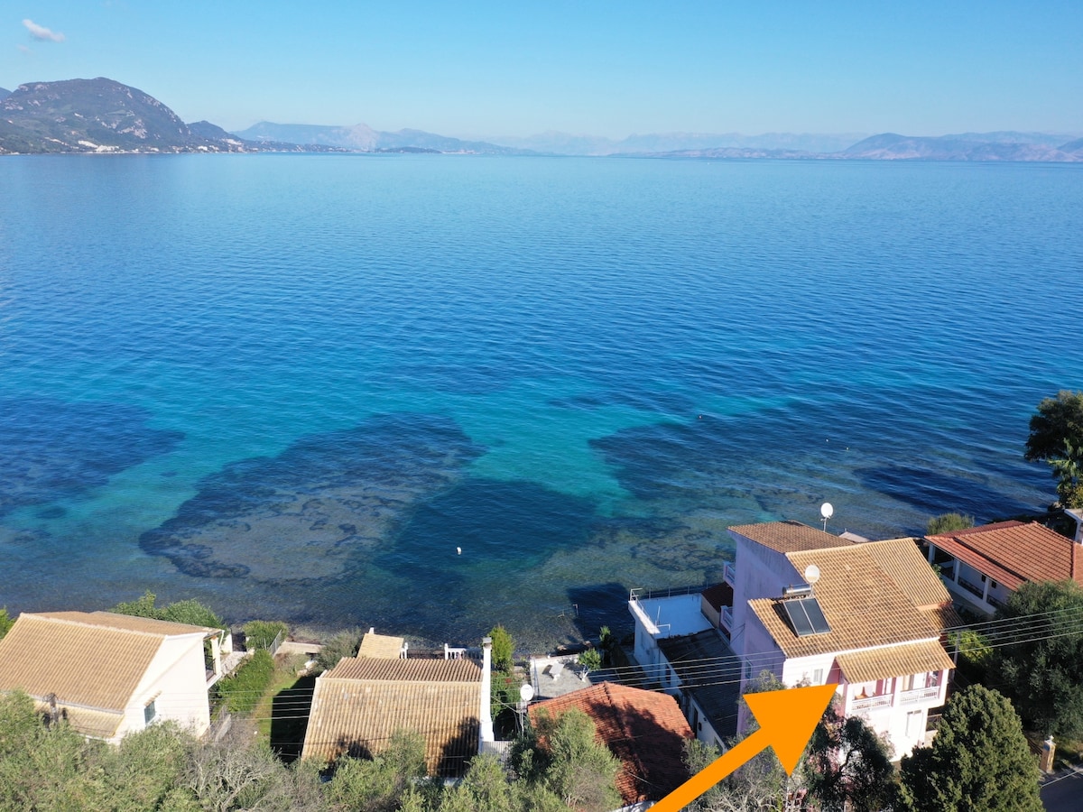 An aerial view showcases a coastal landscape with tranquil blue waters extending to the horizon. The building, indicated by an arrow, features a red-tiled roof and is surrounded by greenery. The calm sea reflects the clear sky, enhancing the serene atmosphere of the setting.