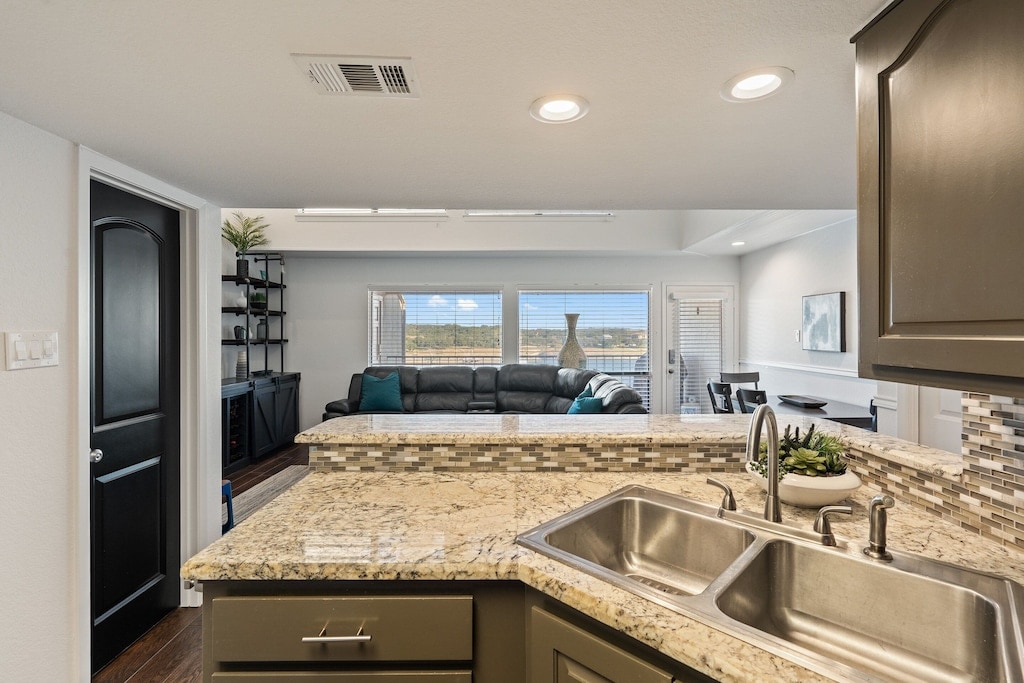 A modern kitchen area is displayed with a textured granite countertop and dual sinks. Beyond the kitchen, a spacious living area is visible, featuring a dark sectional couch and large windows that offer natural light and views of the outdoor scenery.