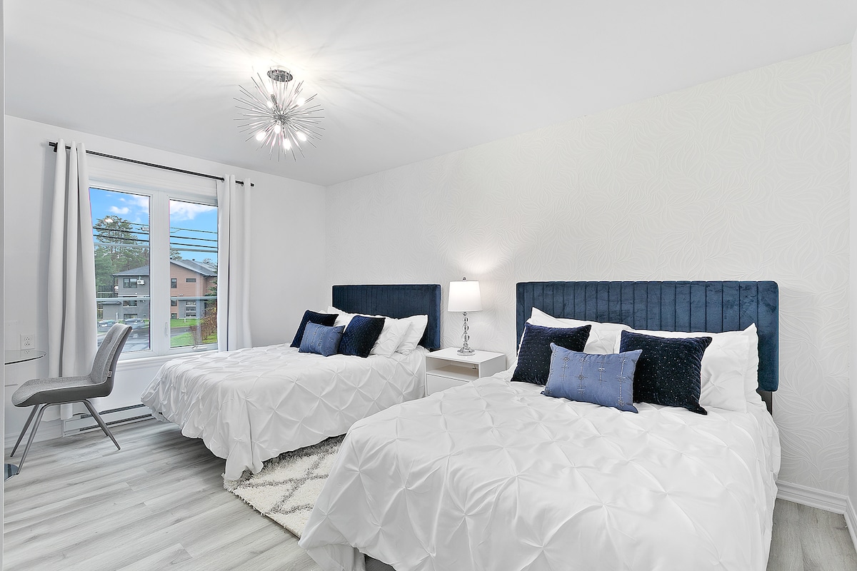 The room features two spacious queen beds with sleek navy headboards and light bedding. A modern light fixture hangs from the ceiling, while natural light streams through a large window dressed with white curtains, illuminating the neutral-toned decor.