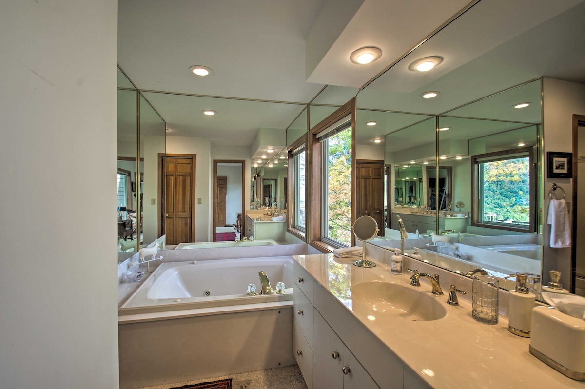 A spacious bathroom is featured, highlighting a large jetted tub next to a wide, mirrored vanity with polished fixtures. Natural light is provided by ample windows, offering views of the surrounding nature. Soft, neutral colors enhance the serene atmosphere.