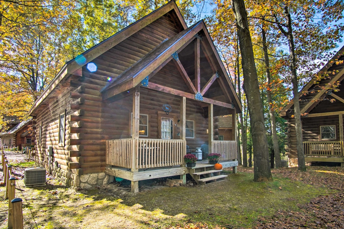 A rustic cabin features a spacious front porch with wooden railings and seating, set amidst vibrant autumn foliage. Wooden logs construct the exterior, showcasing a charming triangle-shaped roof. Sunlight filters through the trees, enhancing the inviting outdoor space.