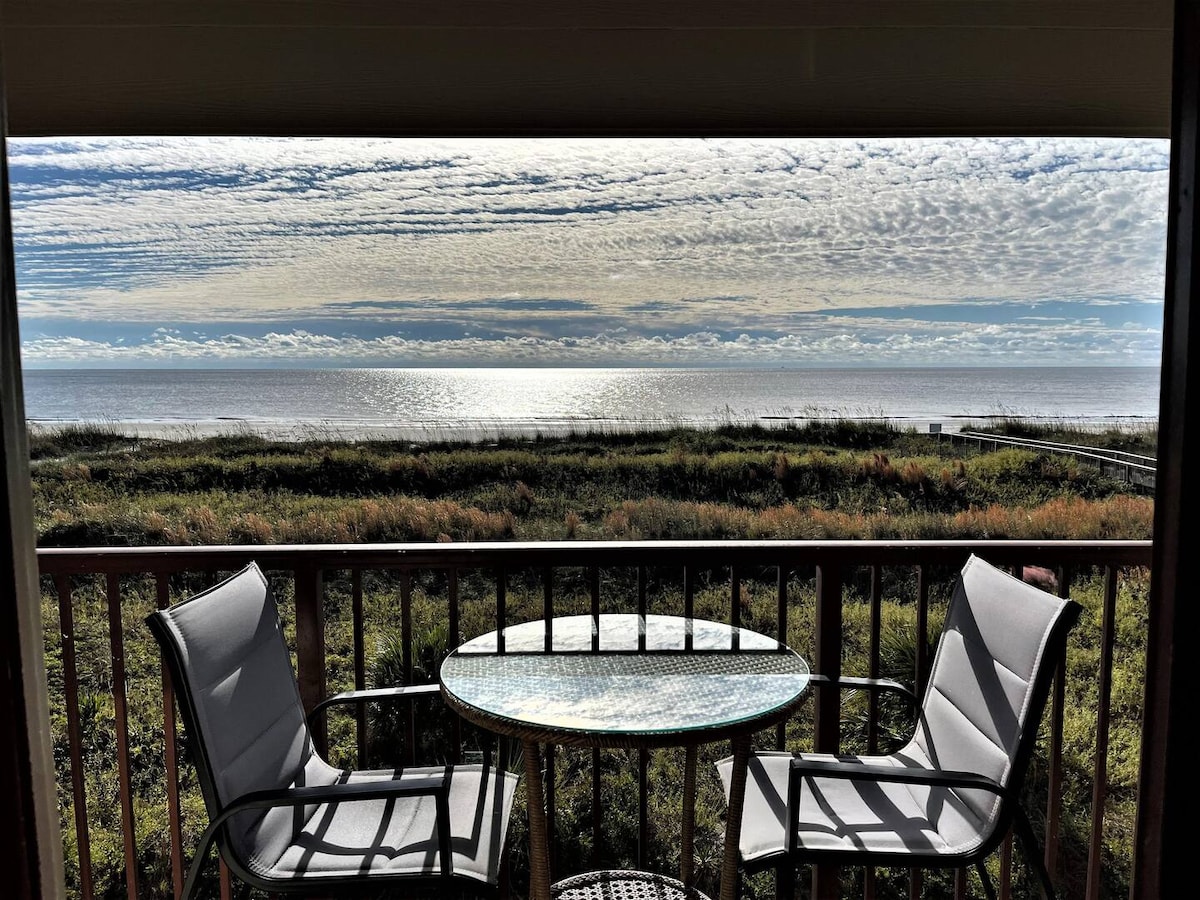 A balcony view features a round glass-topped table surrounded by two cushioned chairs, overlooking a serene ocean scene. The horizon blends gently into the sky, displaying a soft array of clouds reflecting the morning light over calm water.