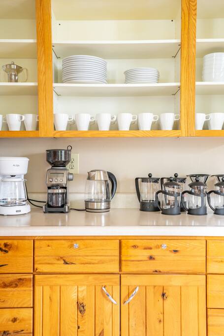 A kitchen space features wooden cabinetry and a countertop with multiple coffee makers, including a drip coffee maker, a kettle, and several French presses. Above, white dishes are neatly arranged on shelves, complemented by a glass storage container.