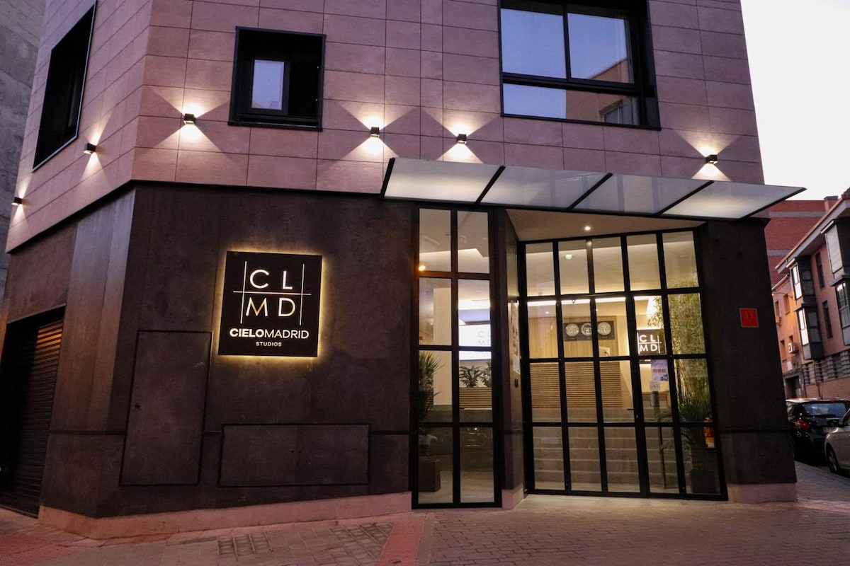 The building entrance is characterized by a sleek, modern façade featuring a combination of stone and dark materials. An illuminated sign displaying 'Cielo Madrid' is positioned prominently, while large glass doors invite guests into the lobby, which is well-lit by soft interior lighting.