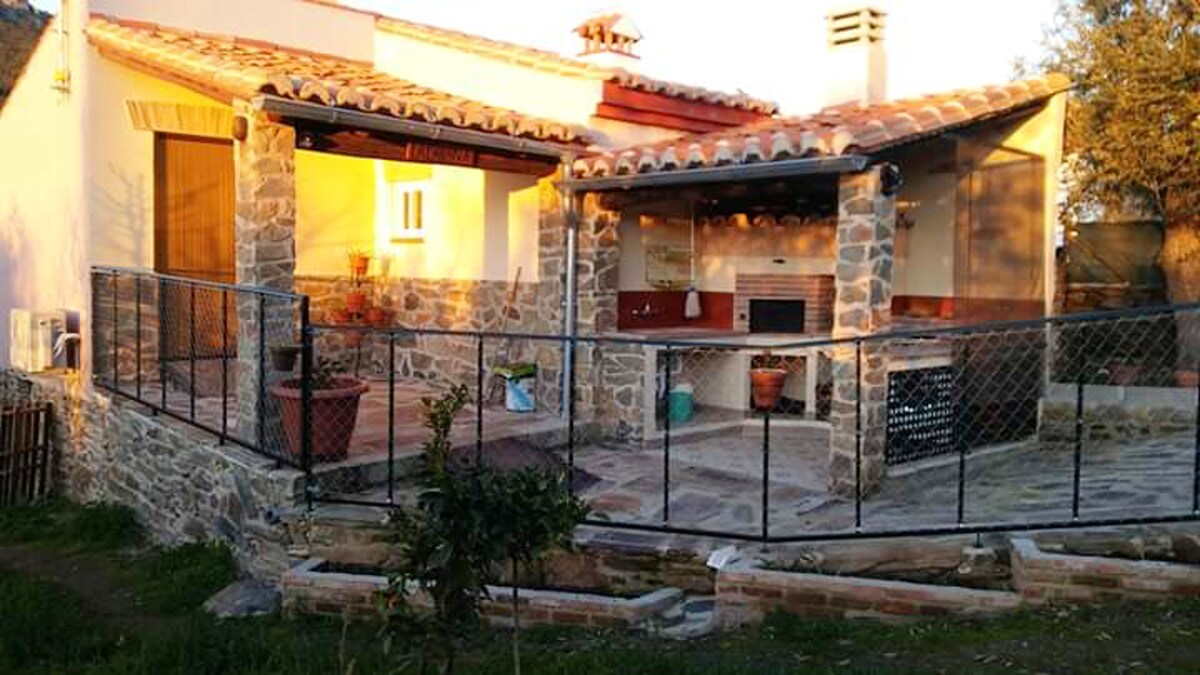 An inviting terrace features outdoor furniture and a built-in grill, surrounded by a fenced garden. Natural stone walls enhance the rustic charm. Sunlight casts a warm glow on the structure, highlighting the cozy outdoor dining area and the surrounding greenery.