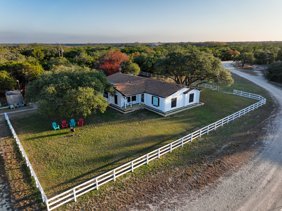 Aerial view of the property reveals a single-story home surrounded by lush greenery on a spacious lot. White fencing encircles the yard, with a cozy seating area featuring colorful chairs. The winding gravel road leads into the peaceful ranch landscape.