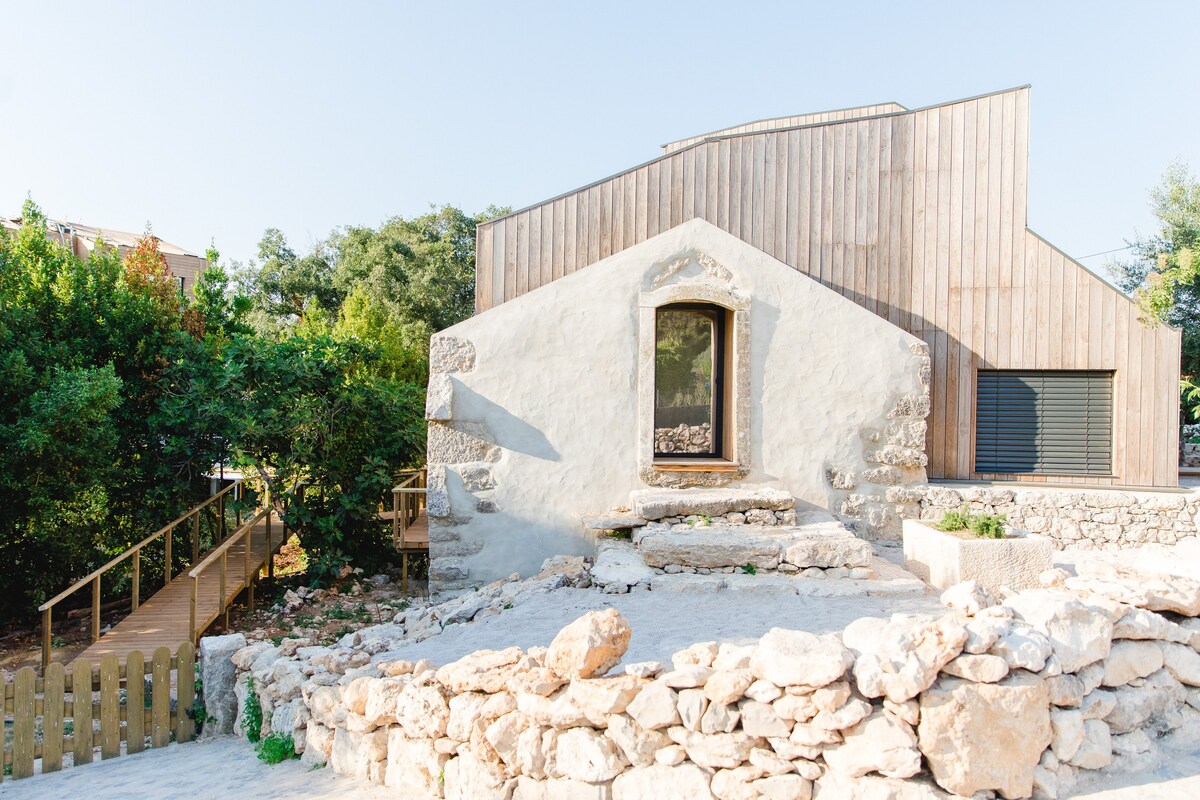 A modern structure featuring a combination of stone and timber. A large window offers a glimpse into the interior. Natural elements, including greenery and a stone pathway, surround the building, enhancing the outdoor space.