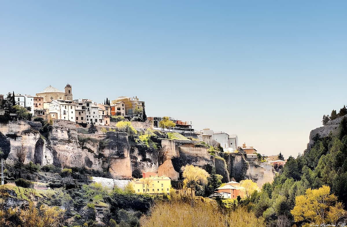 The image captures a panoramic view of Cuenca's picturesque landscape, showcasing multi-colored historical buildings perched on rugged cliffs. The natural surroundings feature lush greenery and trees, while a clear sky spans above, highlighting the architectural beauty of the city.