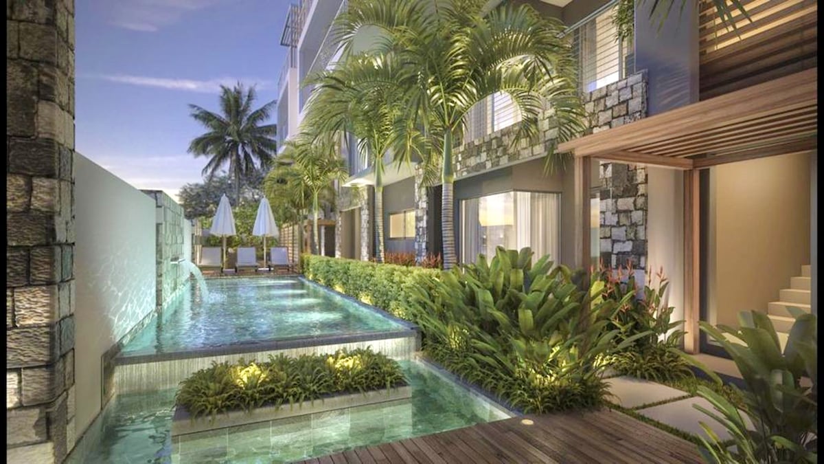 The outdoor area features a shared pool, surrounded by lush greenery and palm trees. Sun loungers are positioned near the pool, offering a space for relaxation. The pool's water reflects the surrounding landscape, complemented by a serene evening sky.
