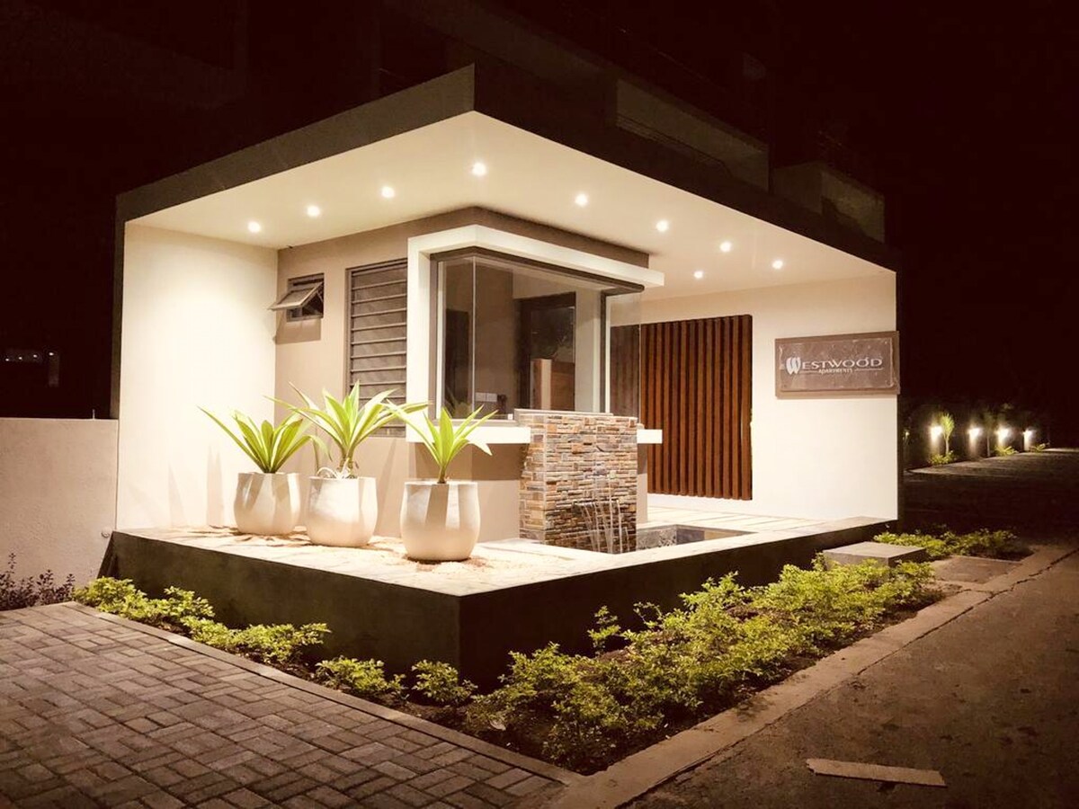 The entrance features a modern façade illuminated by recessed lighting, highlighting planters with leafy green plants. A decorative stone wall complements the entrance area, creating a welcoming ambiance at night.