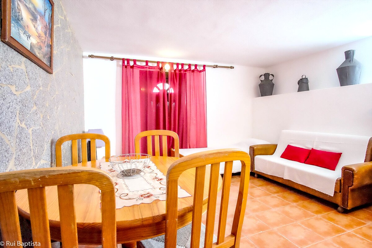 A relaxed dining area is visible, featuring a round wooden table surrounded by several chairs. A soft couch with red accent cushions sits against the wall, and a vibrant curtain partially covers a window. Decorative items are displayed on shelves in the background.