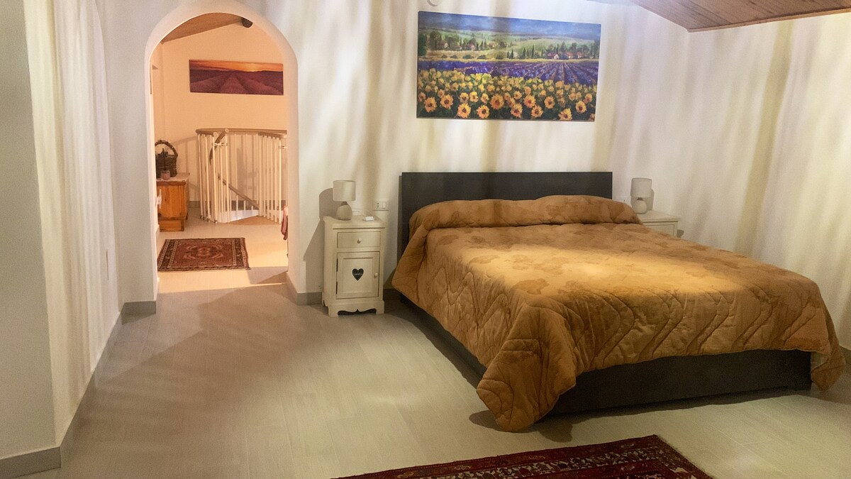 A spacious bedroom features a large bed with a golden-brown quilt. Two nightstands with lamps are positioned on either side of the bed. A piece of artwork depicting sunflowers hangs above the bed, while warm light filters through the room, creating soft shadows.