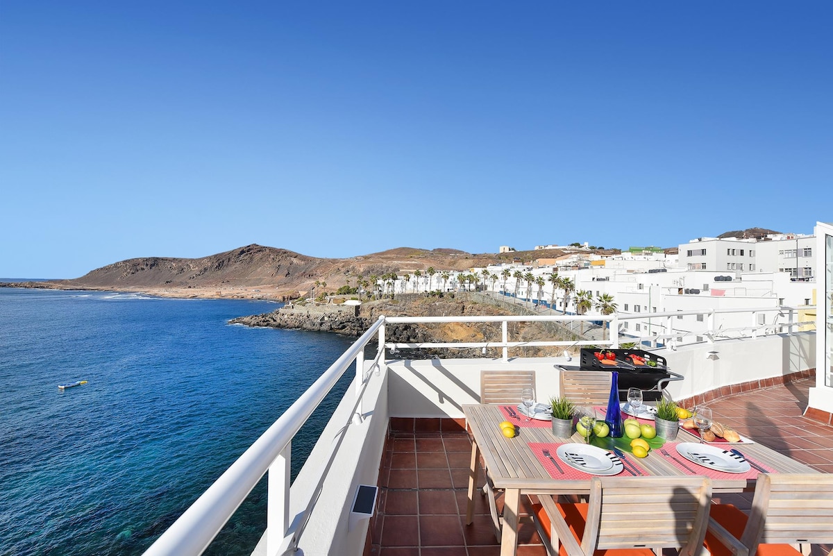A spacious rooftop terrace overlooks the ocean and features a dining table set for outdoor meals. A barbecue grill is positioned nearby, with fruits and decorative items placed on the table. Scenic coastal views and clear blue skies enhance the inviting atmosphere.